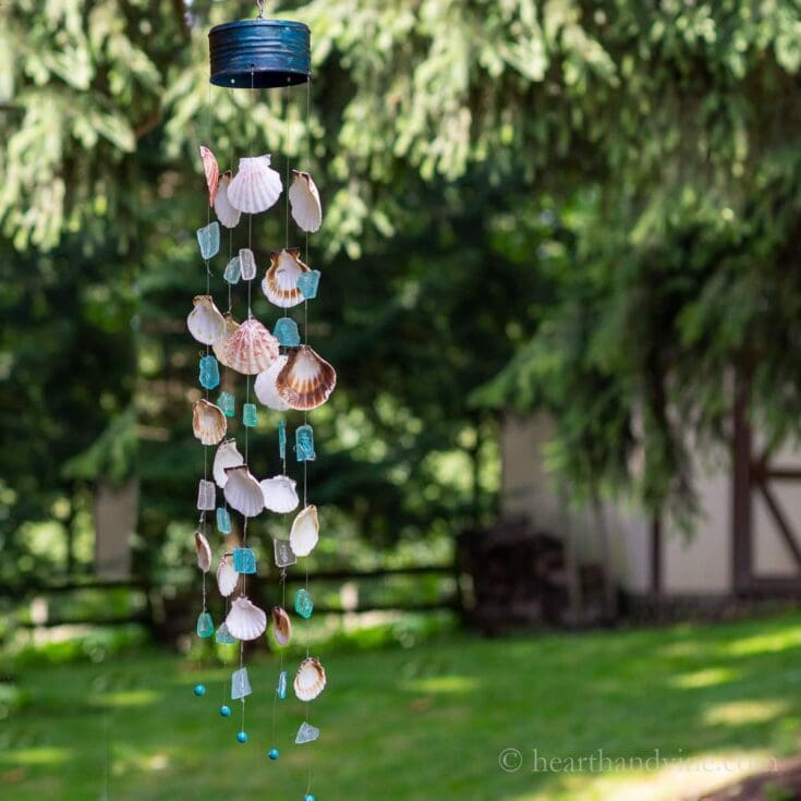 Sea shell and blue glass wind chime hanging outdoors with lush greenery background, creating a charming, coastal-inspired decorative piece for home or garden.