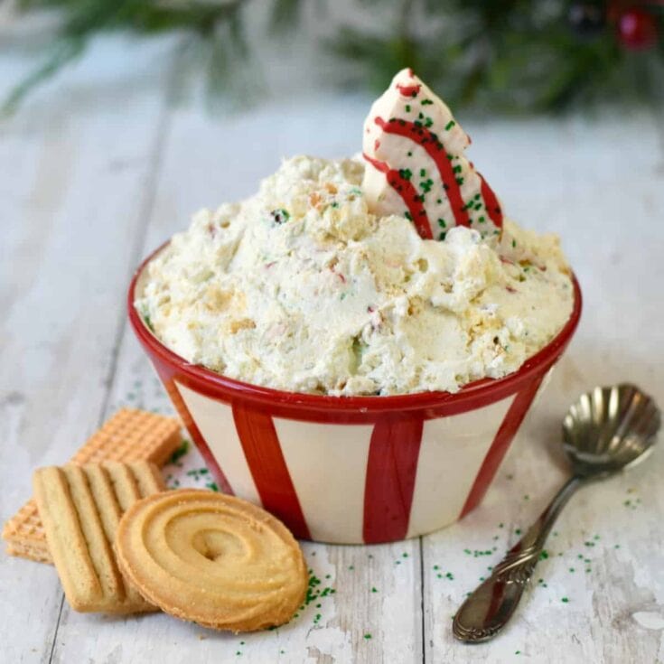 Creamy Christmas-themed cookie ice cream in a red and white striped bowl, topped with holiday sprinkles and a festive cookie, surrounded by assorted cookies on a rustic white surface.