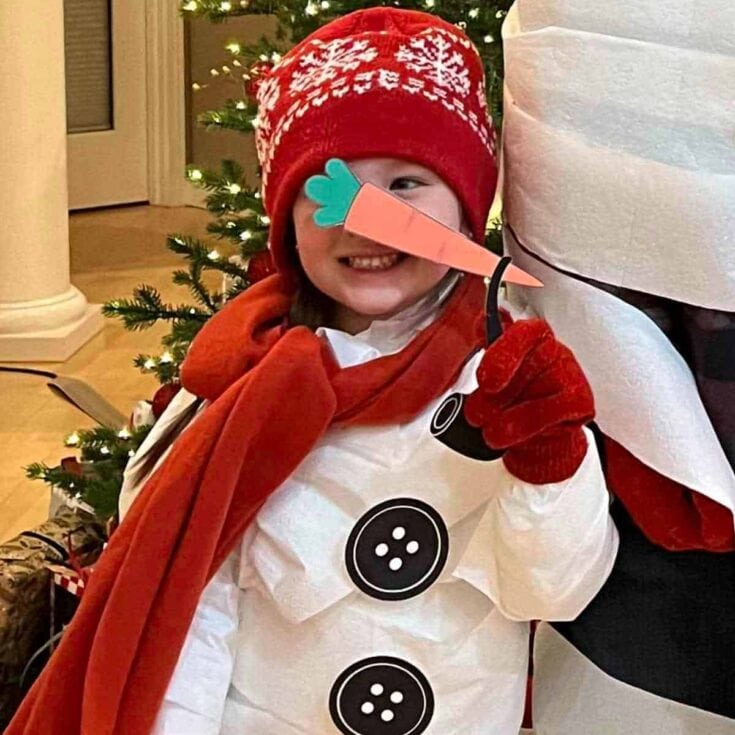Cute child dressed as a snowman with a carrot nose, red winter hat, and festive holiday attire, smiling happily during Christmas celebrations near a decorated tree.