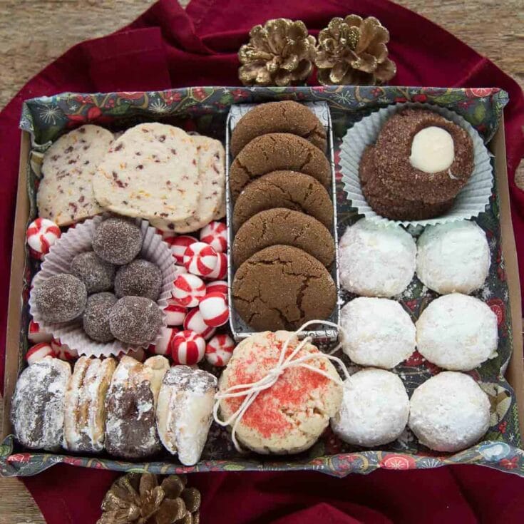 Gingerbread cookies, peppermint candies, chocolate truffles, and assorted holiday cookies in a festive gift box on red fabric with pinecones and holiday decorations.