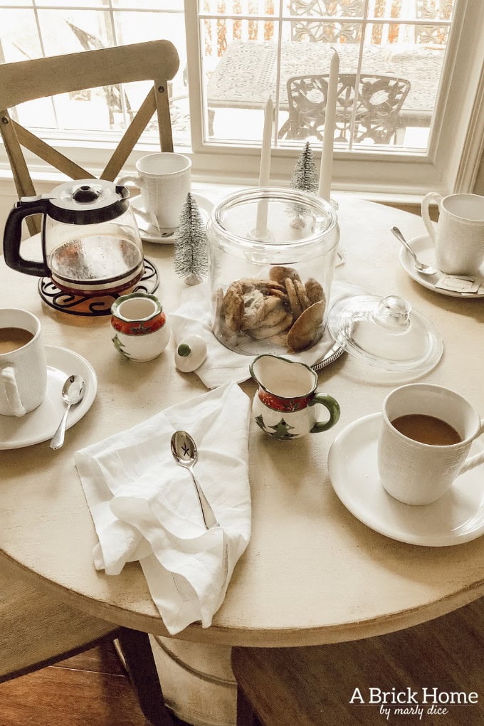 A round table set for breakfast with teapots, cups, a jar of cookies, and a napkin with a spoon, near a window with a snowy outdoor scene in the background.