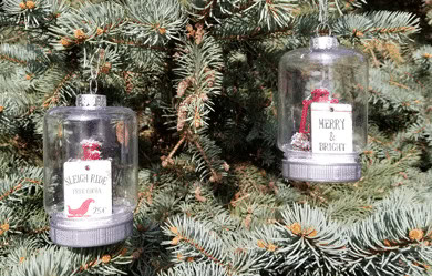 Two glass dome Christmas ornaments with candles and festive messages on a pine tree background, creating a cozy holiday atmosphere.