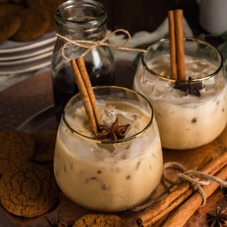 Cinnamon-spiced iced coffee drinks garnished with cinnamon sticks and star anise, served in elegant glasses on a rustic tray, with cookies and a glass jar in the background.