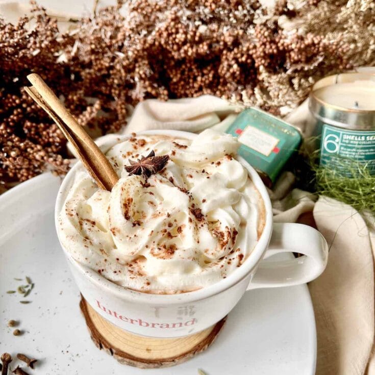 Warm creamy hot chocolate topped with whipped cream, cinnamon, and star anise, served in a branded mug with cozy herbal and candle accents for a comforting beverage experience.