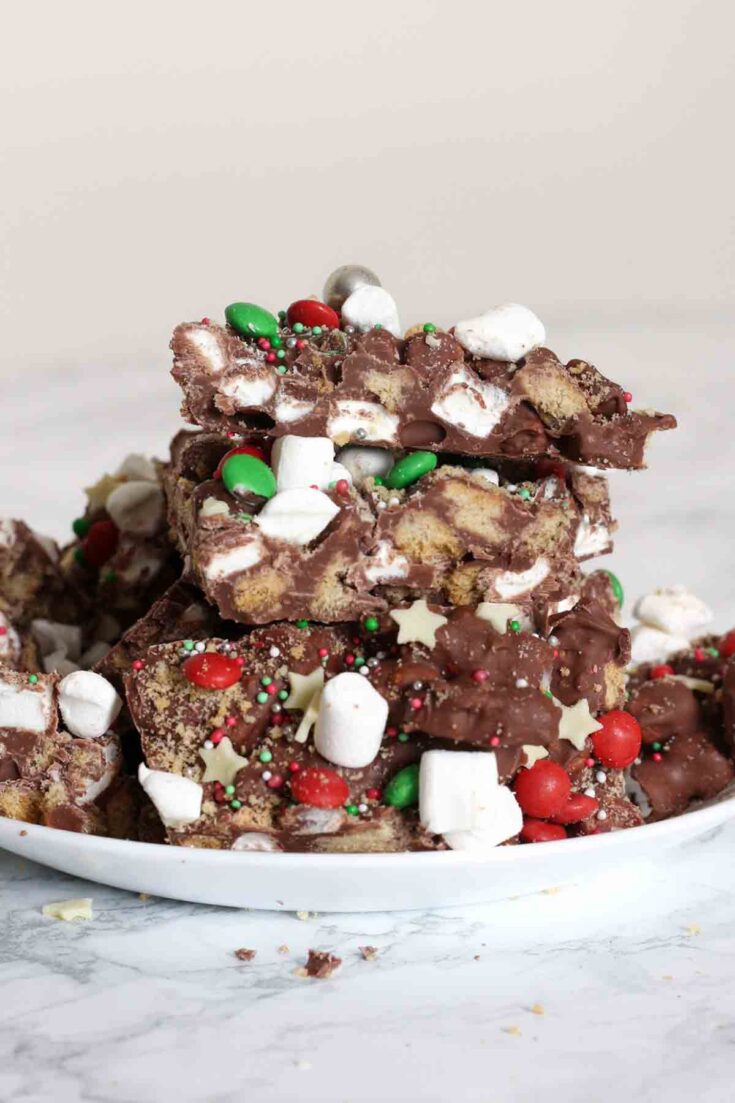 Decadent holiday rocky road bark topped with colorful holiday sprinkles, mini marshmallows, and chocolate candies on a white plate, perfect for festive Christmas treats.