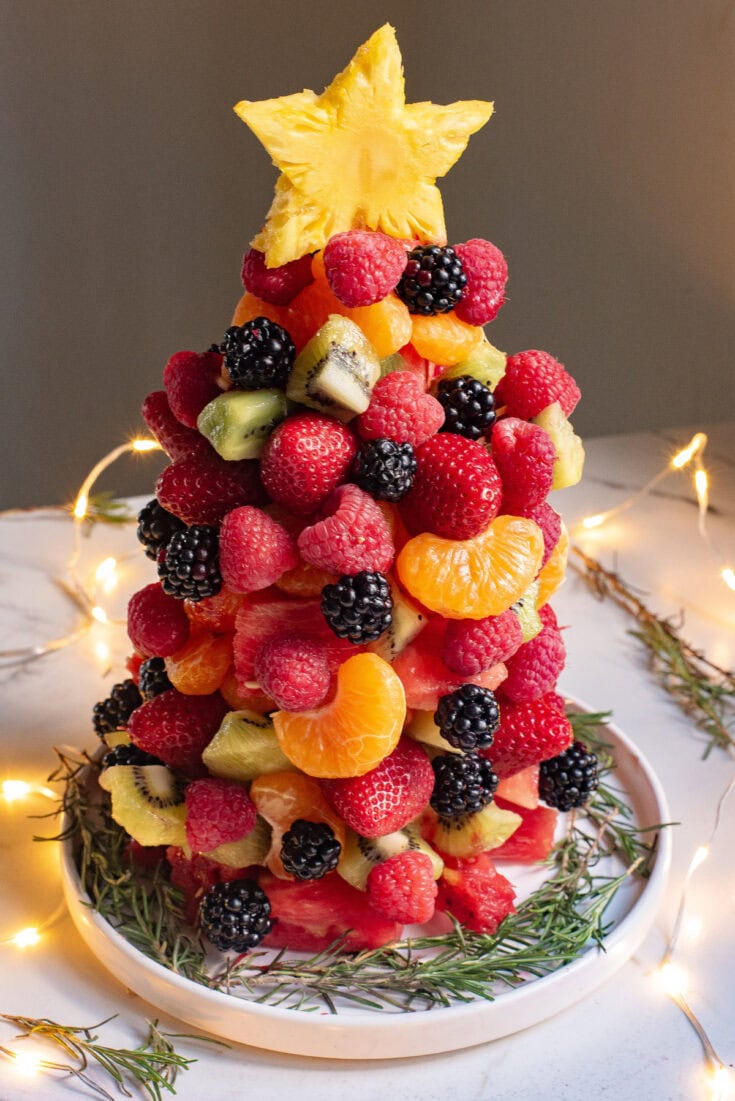 Colorful fruit Christmas tree decorated with strawberries, raspberries, blackberries, kiwi, pineapple, and mandarin oranges, on a white platter, surrounded by fairy lights for festive holiday celebration.