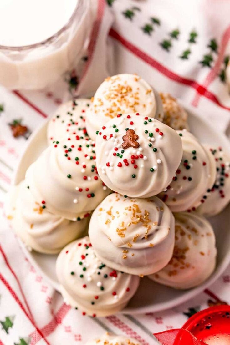 Festive holiday cookies with white icing, colorful sprinkles, and gingerbread decor, arranged on a plate with a Christmas-themed cloth in the background. Perfect for Christmas celebrations.