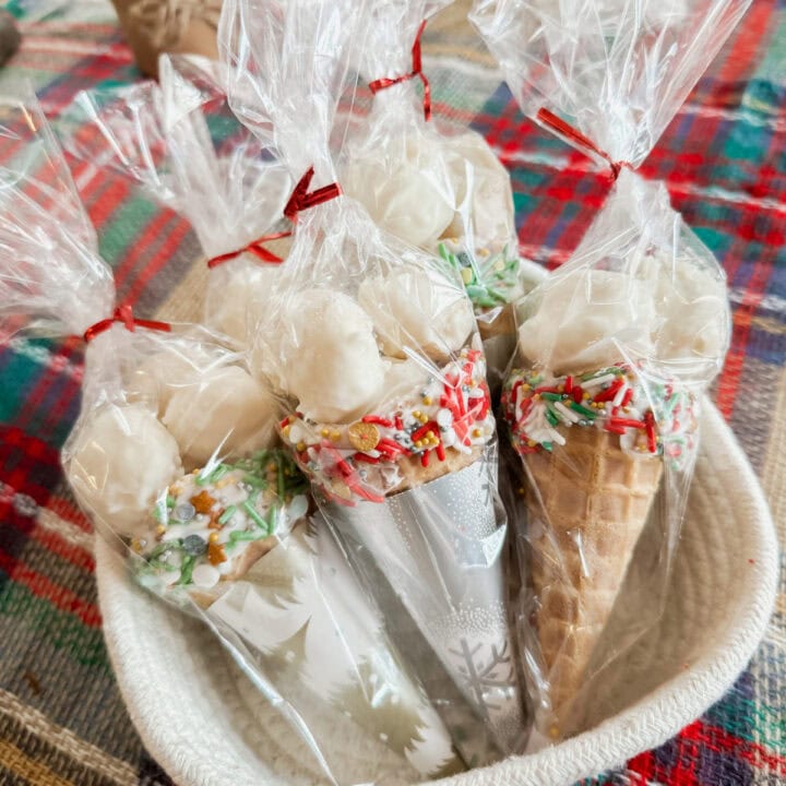 Hand-decorated holiday ice cream cones with festive sprinkles, wrapped in clear plastic, placed in a white basket on a colorful plaid tablecloth. Perfect for Christmas desserts or winter celebrations.