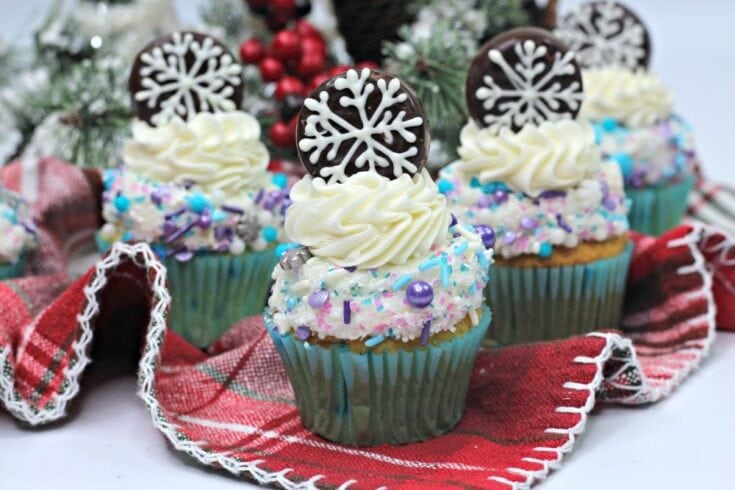 Colorful Christmas cupcakes decorated with snowflake chocolate toppers, sprinkles, and whipped frosting on a festive red and white napkin. Perfect for holiday celebrations.