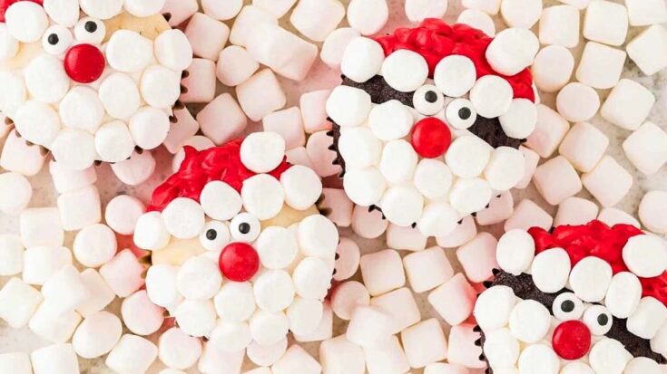 Festive Santa-themed cupcakes decorated with marshmallows, red icing, and candy eyes, surrounded by mini marshmallows, perfect for Christmas celebrating with adorable holiday treats.