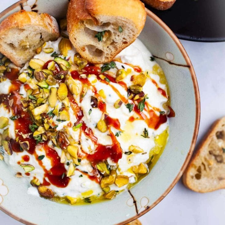 Creamy Greek yogurt dip topped with chopped pistachios, fresh herbs, and hot sauce served with toasted baguette slices, perfect for appetizer or snack.
