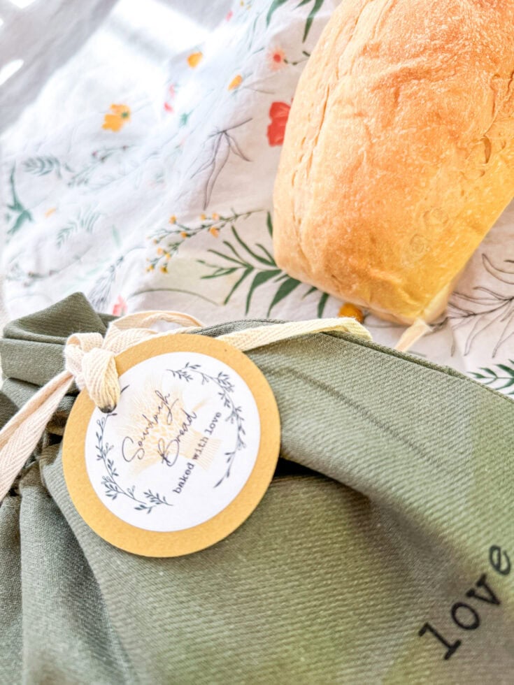 Lavender baked bread with a tag reading "Sunday Bread" on a green cloth bag, set on a floral tablecloth, showcasing artisanal, homemade baked goods perfect for gifting or breakfast.