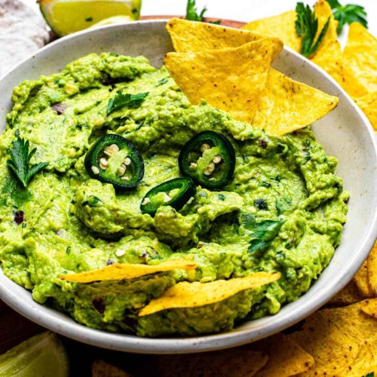 Creamy guacamole dip with jalapeño slices and fresh herbs, served with crispy tortilla chips and lemon wedges, perfect for snacking and party appetizers.