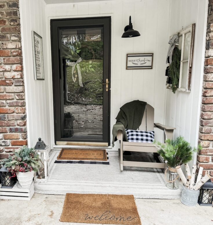 Cozy farmhouse front porch decorated with Christmas greenery, black and white accents, and inviting welcome mat, creating a warm holiday entryway scene.