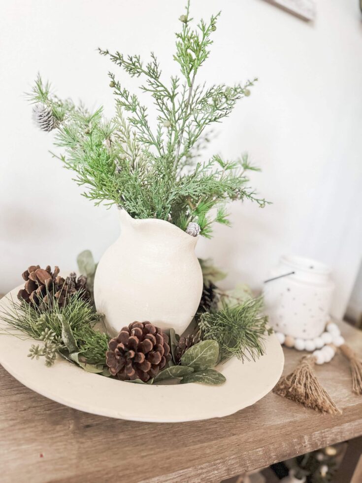 Minimalist holiday centerpiece with greenery and pinecones in a white ceramic vase on rustic wooden surface.
