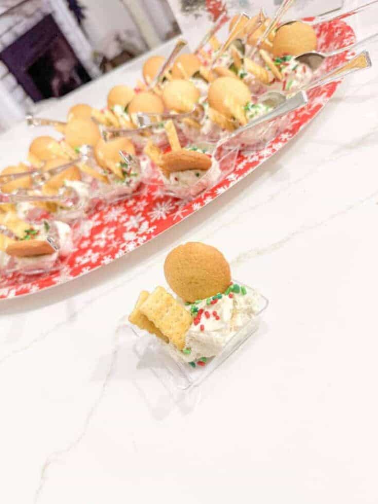 Mini holiday cookies and festive whipped cream dessert on a marble countertop with a holiday-themed tray of assorted treats in the background.
