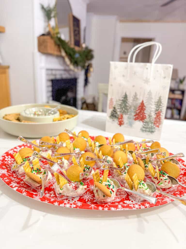 Festive holiday appetizer tray with finger foods, cookies, and a holiday-themed gift bag on a white table, celebrating Christmas in a cozy home setting with a fireplace and decorations.
