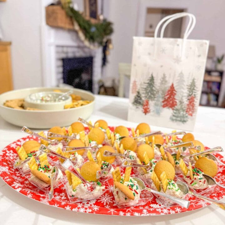 Festive holiday appetizer tray with finger foods, cookies, and a holiday-themed gift bag on a white table, celebrating Christmas in a cozy home setting with a fireplace and decorations.