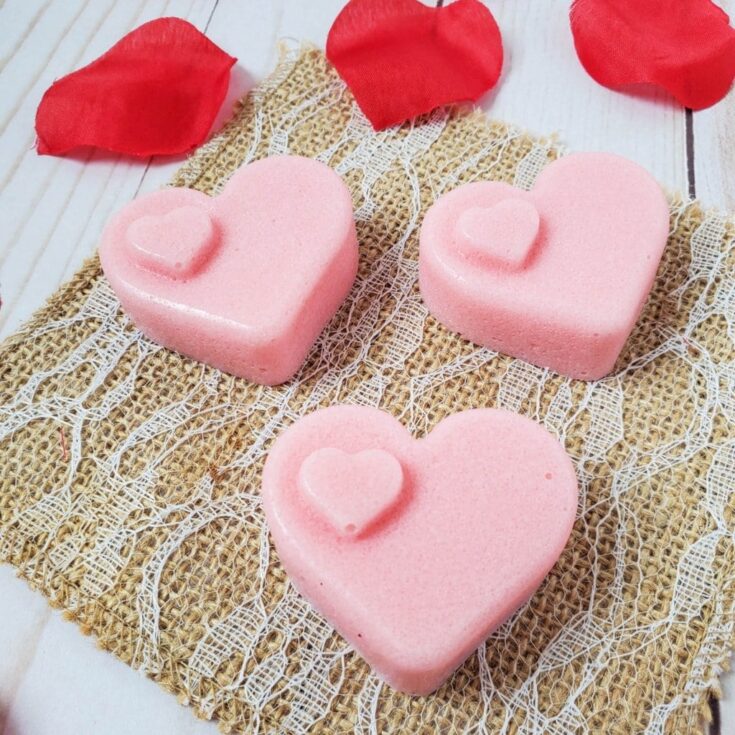 Pink heart-shaped bath bombs with small heart accents, surrounded by red rose petals on a rustic textured surface, representing love and self-care.