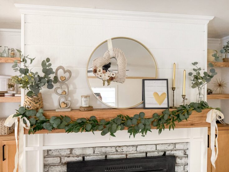 Eucalyptus garland decorated on a wooden mantel over a white brick fireplace, with a round gold-framed mirror and framed heart artwork, candles, and decorative items creating a cozy farmhouse aesthetic.