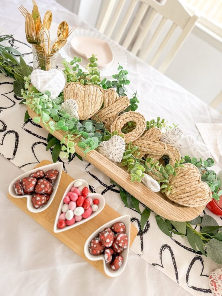 Wicker heart-shaped decor and greenery centerpiece with candy dishes for romantic Valentine's Day table setting.