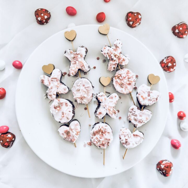 Sweetheart cookie pops with pink icing and sprinkles on a white plate, surrounded by red and white heart-shaped chocolates and candies, perfect for Valentine's Day celebrations.