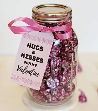 Colorful Valentine's Day gift jar filled with pink foil-wrapped chocolates, decorated with a pink ribbon and a "Hugs & Kisses" tag, perfect for expressing love and affection.