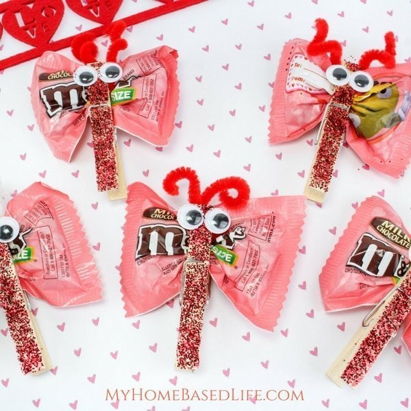 Pink Valentine-themed homemade gift decorations featuring M&M's chocolate wrappers styled as cute bugs with googly eyes, red pipe cleaner antennae, and glittery candy stick legs, perfect for romantic or love celebrations.
