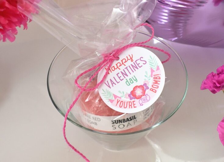 Pink Valentine's Day soap with "Happy Valentine's Day You're the Bomb!" gift tag in a glass bowl, surrounded by flowers.
