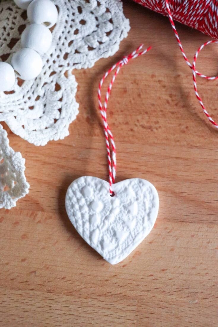 Delicate white heart-shaped ornament with intricate textured design, hanging on red and white twine, surrounded by crochet lace and festive decor on a wooden surface for Christmas or holiday gifting.