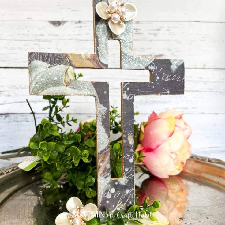 A distressed metal cross with pearl and floral embellishments, surrounded by greenery and a pink peony, on a rustic white wooden background, perfect for religious or spiritual decor.