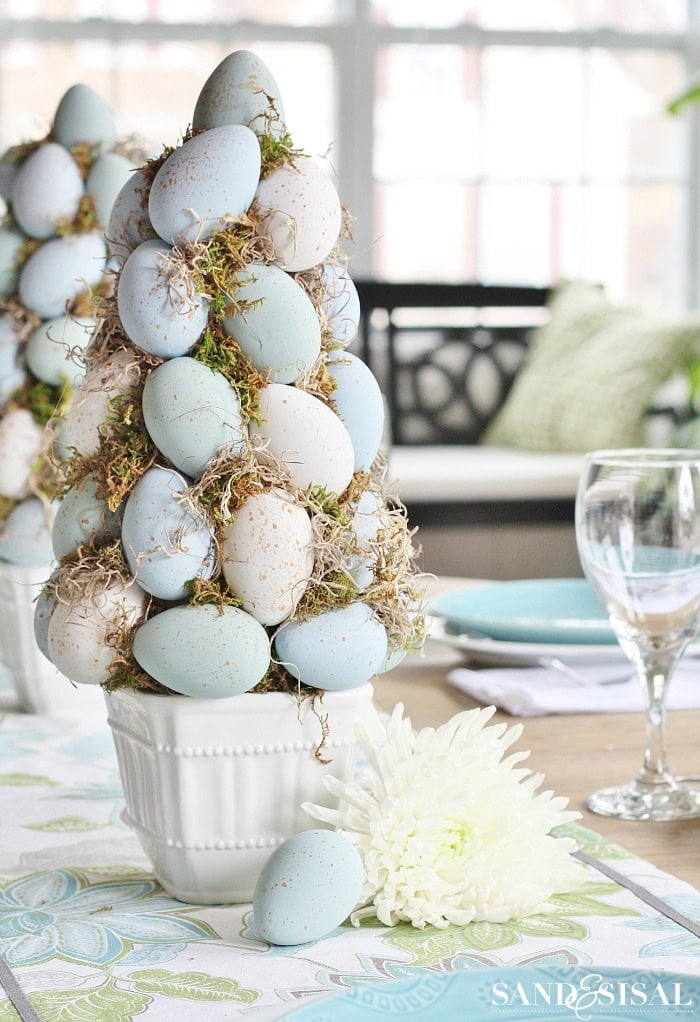 Egg centerpiece with pastel blue and white eggs stacked in a white ceramic pot, decorated with moss and twigs, placed on a table with floral tablecloth and elegant table setting.
