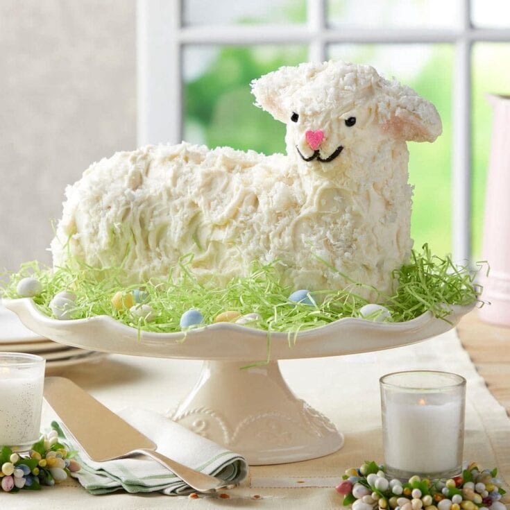 Lamb-shaped cake decorated with white frosting and edible details, displayed on a cake stand with pastel Easter eggs and greenery, perfect for spring celebrations.