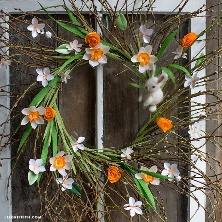 Delicate spring floral wreath with white and orange daffodils, greenery, bunny ornament, on rustic wooden background, perfect for Easter or spring decor.