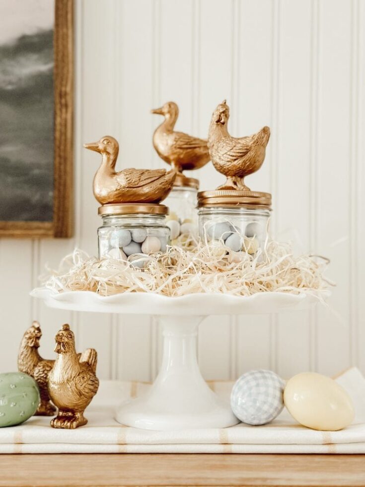 Golden duck figurines and Easter eggs displayed on a white cake stand for spring and Easter celebrations.