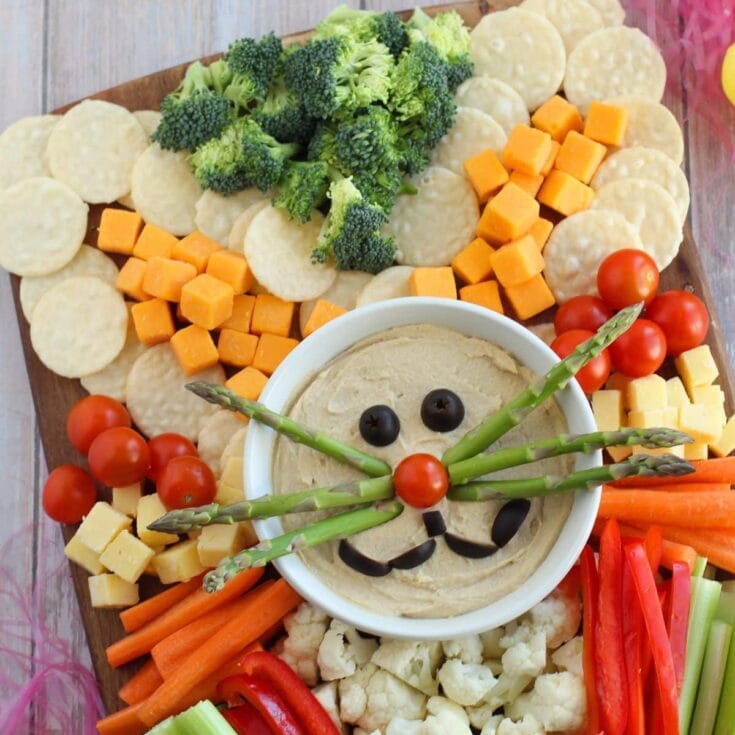 Colorful vegetable platter with hummus dip decorated as a friendly snowman, featuring cherry tomatoes, broccoli, cheese cubes, carrots, cauliflower, and celery sticks. Perfect for festive or healthy snacks.