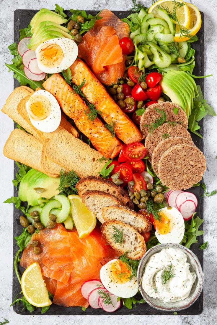 Colorful smoked salmon and veggie platter with boiled eggs, avocado, cherry tomatoes, cucumber ribbons, rye bread slices, capers, lemon wedges, and flavored cream cheese.