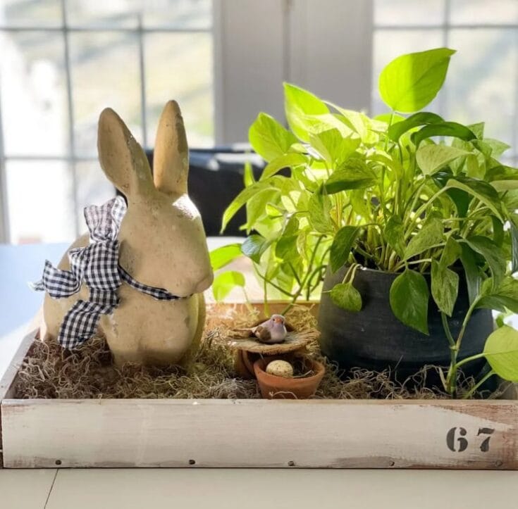 Handcrafted ceramic bunny with a gingham ribbon, surrounded by a lush green potted plant, a small bird figurine, and nest accents in a charming spring-themed decorative tray.