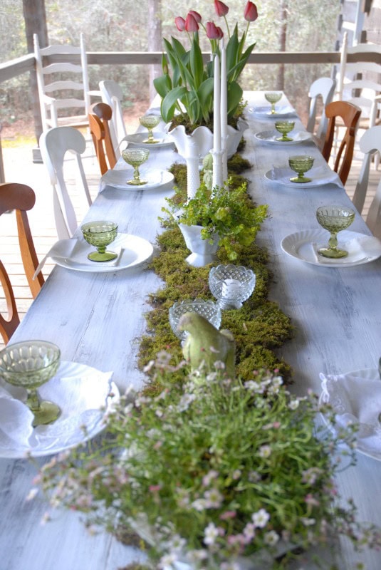 Elegant spring table setting with fresh pink tulips, moss centerpiece, vintage green glassware, and white dishes on a sunlit porch for outdoor dining and celebration.