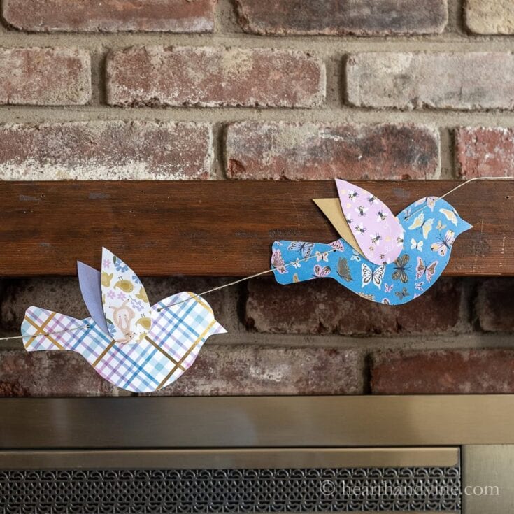 Paper bird garland with colorful patterned designs hanging on a wooden ledge against a red brick wall.