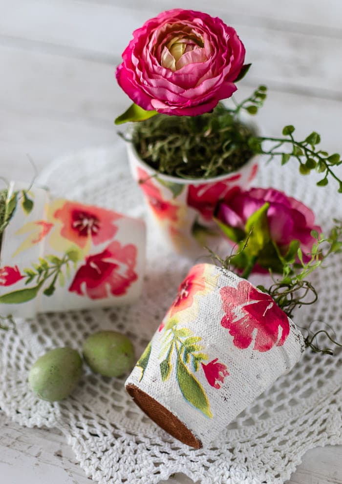 Pink ranunculus flower in a decorative floral cup with watercolor floral design, surrounded by small green fruits and additional floral cups on a white textured surface, emphasizing handcrafted floral decor and home accents.