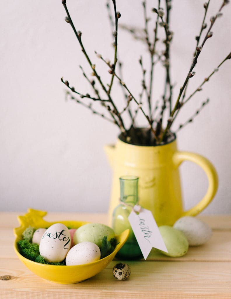 Elegant yellow vase with blooming branches and decorated Easter eggs on a wooden table.