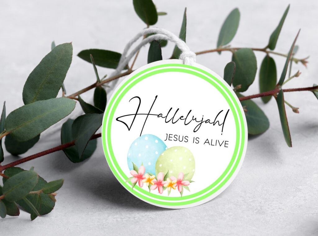 Easter egg sticker with "Hallelujah! Jesus is alive" message, surrounded by pastel-colored eggs and pink flowers, on a sprig of green eucalyptus leaves.