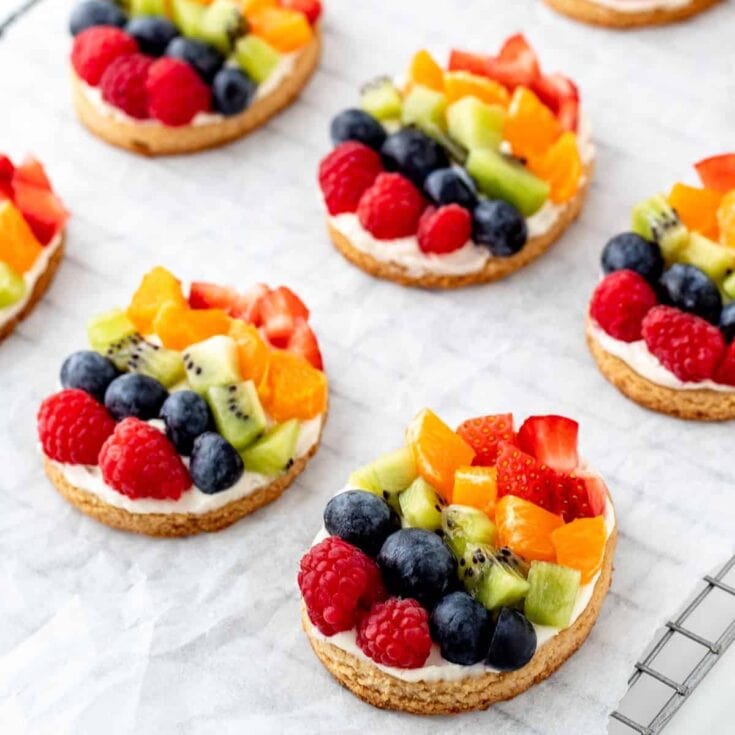 Fresh fruit tart cookies topped with blueberries, raspberries, strawberries, kiwi, and oranges, on a white parchment paper background, showcasing colorful, healthy, and delicious treat options.