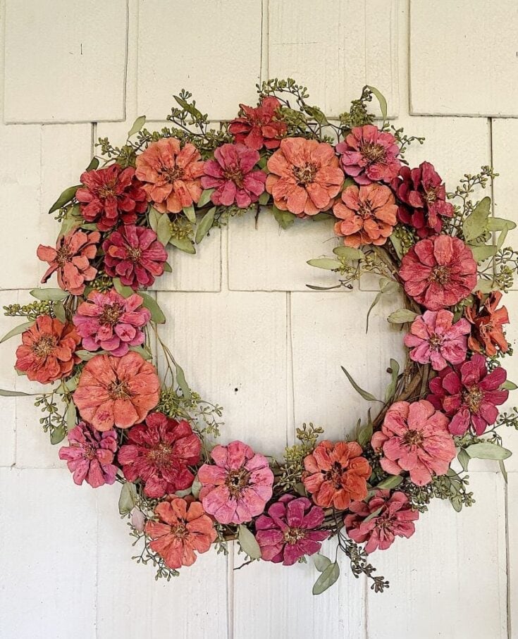 Colorful floral wreath with pink, peach, and red blooms arranged in a circular shape on a white wooden background. Perfect for spring or summer decor, weddings, or special occasions.