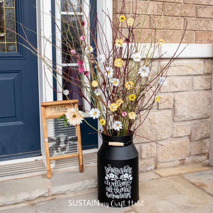 Colorful spring flower arrangement in a black vase with inspirational quote, placed on a porch in front of a blue door and brick wall, perfect for home decor and seasonal decorations.