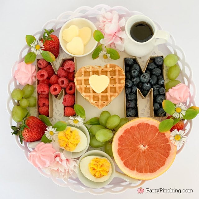 Fresh fruit and waffle breakfast platter spelling "MOM" with strawberries, blueberries, green grapes, a halved grapefruit, sliced boiled eggs, a small heart-shaped waffle, white chocolate hearts, pink flowers, and black coffee on a white decorative tray.