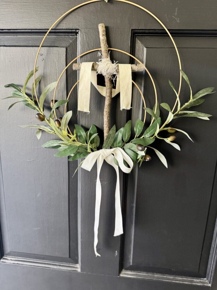 Rustic olive branch wreath with wood cross and fabric ribbons hanging on a black door, perfect for religious or holiday decor.