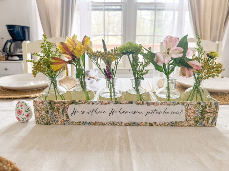 Colorful flower vases with fresh blooms on a bright dining table, decorated with a floral greeting card that reads, "He is not here. He has risen, just as he said," perfect for an Easter or spring celebration.