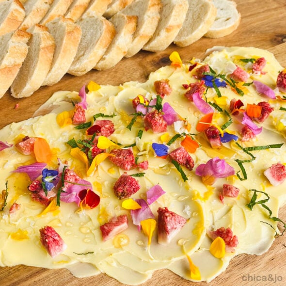 Delicious white chocolate bark topped with colorful edible flower petals, dried berries, and drizzled with white chocolate, served with slices of fresh bread on a wooden cutting board.